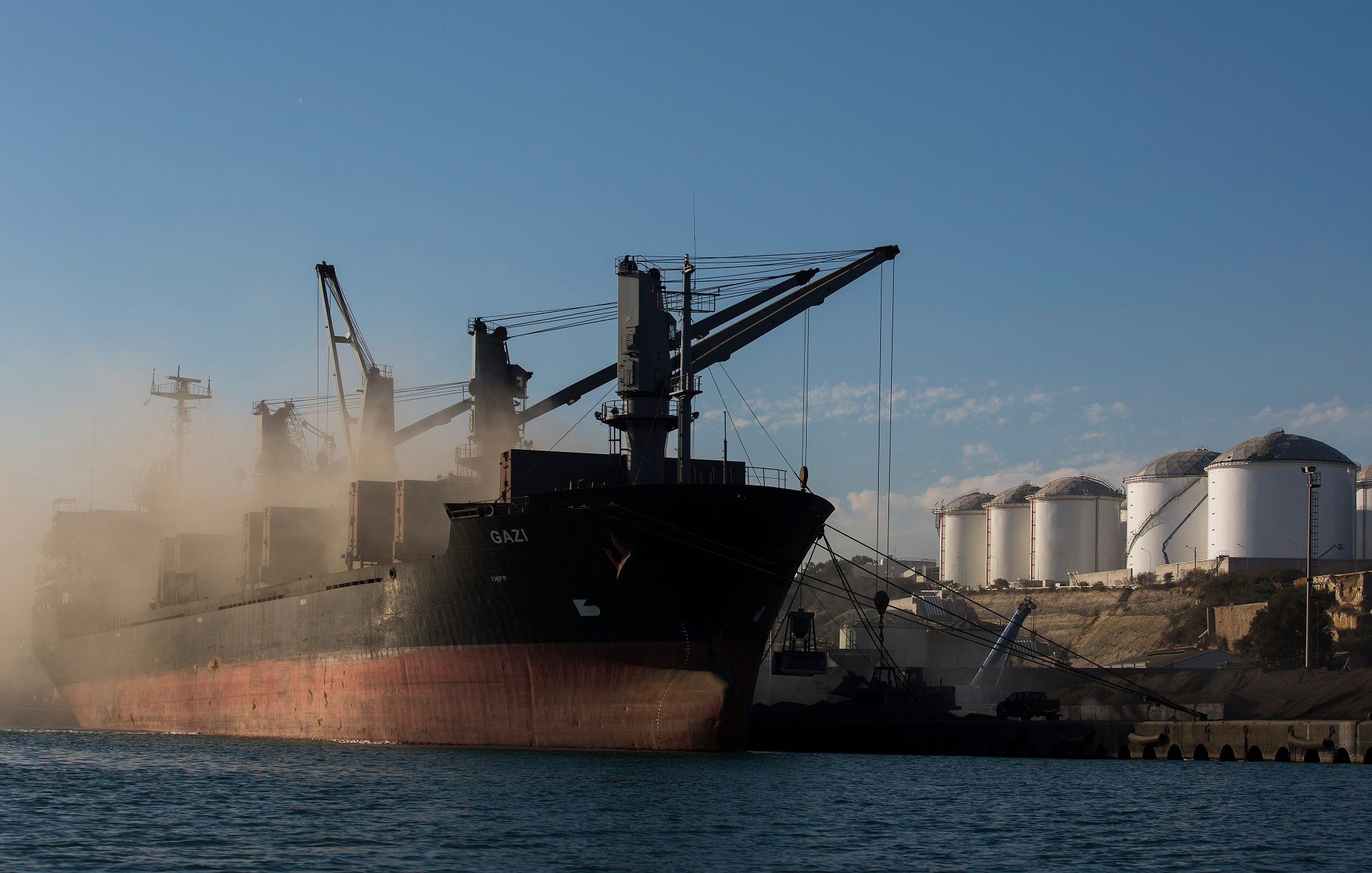 Cement is loaded onto a cargo ship as fuel storage tanks stand on the shore at the VTTV oil storage terminal, a joint venture of Vitol Group and MISC Bhd., in Vasilikos, Cyprus, on Friday, Feb. 21, 2014. Cyprus's economic outlook remains subject to "significant risk" even after its economy contracted less than expected last year, debt monitors said at the end of a quarterly review of its adjustment program.